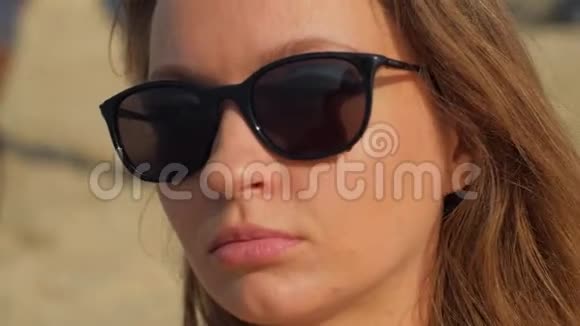 阳光明媚的夏日带着黑色太阳镜在海滩上微笑着的金发年轻女子视频的预览图