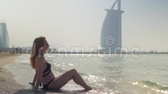 年轻的金发女人穿着黑色比基尼坐在迪拜的沙滩上视频的预览图