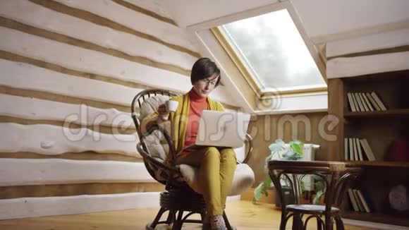 戴眼镜的成年美女在笔记本电脑上看电影在阁楼上喝咖啡视频的预览图