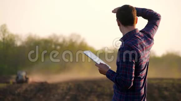 年轻的农民在田间拖拉机前拿着平板电脑农夫用平板电脑控制着田里工作视频的预览图