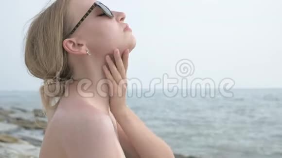 阳光明媚的夏天一个戴太阳镜脖子上涂防晒霜坐在海滨的迷人少女视频的预览图