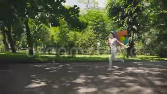 快乐的小女孩在阳光明媚的绿色公园里奔跑手里拿着五颜六色的气球视频的预览图