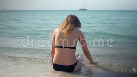 年轻女孩穿着黑色比基尼在阳光明媚的日子里坐在沙滩上后景视频的预览图