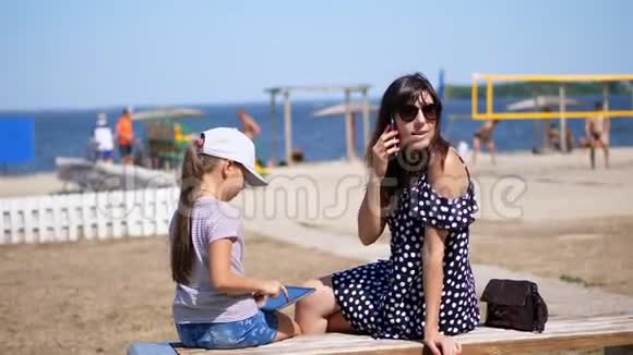 母亲一个戴着太阳镜的漂亮女人带着一个八岁的女儿正坐在炎热的夏日海滩上视频的预览图