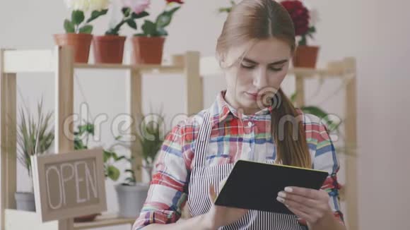 小生意一个穿着格子衬衫和条纹围裙的女孩站在那里手里拿着一块数字平板电脑并计划着看上去视频的预览图