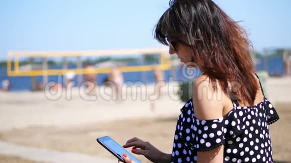 肖像美丽的女孩戴着太阳镜在平板电脑上工作在海滩上在炎热的夏天在一个背景下工作视频的预览图