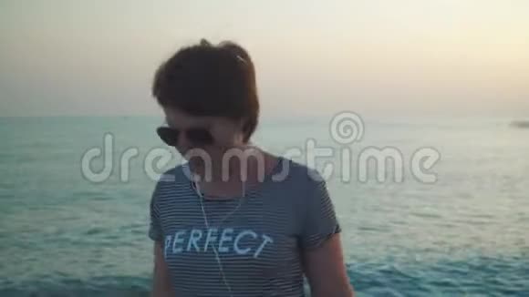 穿着t恤的女人戴着耳机听音乐在夕阳下的海景中悠闲地跳舞视频的预览图