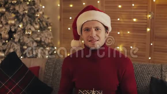 圣诞老人戴着圣诞老人的帽子看着圣诞树背景上的摄像机的微笑迷人男人的肖像慢动作视频的预览图