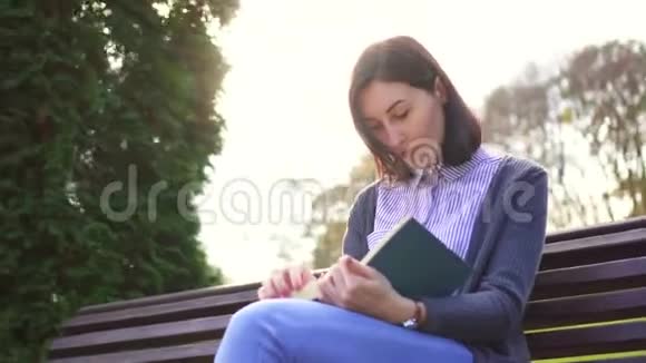 日落时分一个拿着书坐在户外长凳上的女孩视频的预览图