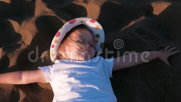 美丽快乐的小女孩金发戴着帽子躺在沙滩上视频的预览图