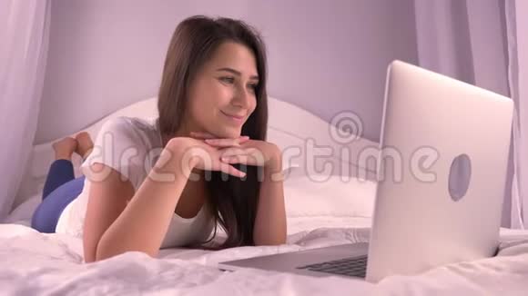 年轻漂亮的少女在笔记本电脑上浏览躺在床上微笑的特写照片视频的预览图