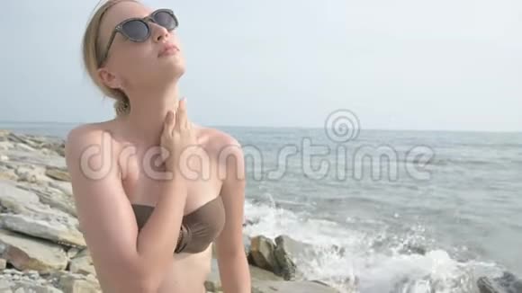 一个年轻迷人的女孩戴着太阳镜脖子和肩膀上涂着防晒霜坐在海边视频的预览图