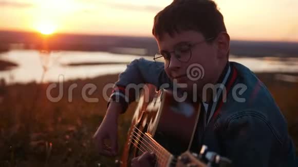 戴着圆眼镜的男孩在日落时分在田野里弹吉他微笑着视频的预览图