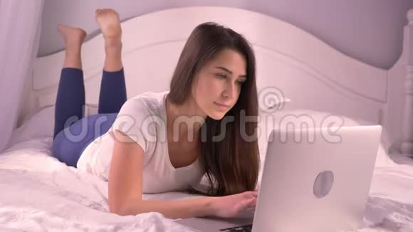 在舒适的家里躺在床上微笑着使用笔记本电脑的年轻漂亮少女的特写照片视频的预览图