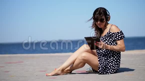美丽的黑发女人戴着太阳镜和耳机的女人在平板电脑上工作听音乐坐在海滩上视频的预览图