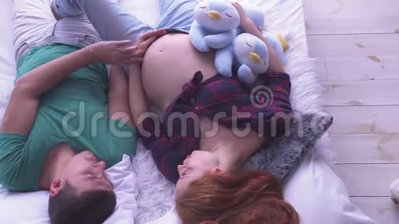 年轻的怀孕夫妇相爱地躺在床上爸爸抚摸妻子的肚子生育观念怀孕视频的预览图