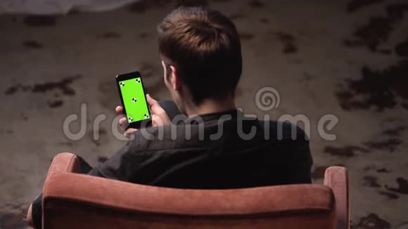 在黑暗的房间里棕色头发的年轻人坐在椅子上敲着iPhone绿色屏幕库存数据视频的预览图
