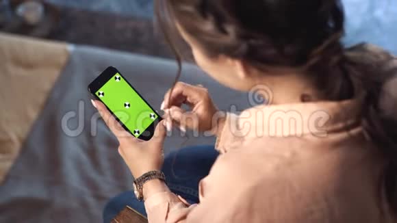 一个棕色头发的漂亮女人穿着粉色衬衫坐在家里在她的绿色屏幕上滑动着她的智能手机视频的预览图