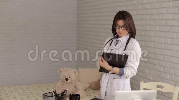 一位戴眼镜的女儿科医生的肖像一件白色长袍和笔记本视频的预览图