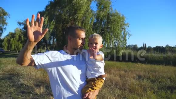 在公园里快乐的父亲和小儿子挥舞着手对着镜头的肖像年轻的爸爸抱着他的孩子视频的预览图