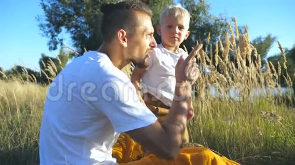 一个阳光明媚的夏日年轻爸爸和他可爱的儿子金发坐在草地上家庭幸福支出视频的预览图