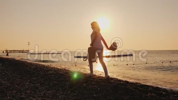 在黎明时分手里拿着背包和帽子的无忧无虑的女人沿着海滨奔跑暑假概念视频的预览图