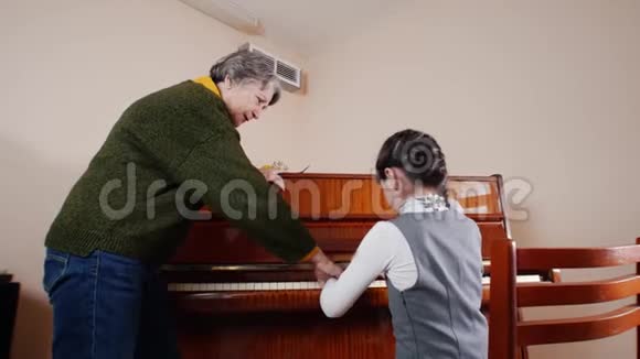 音乐课女孩弹钢琴年长的老师站在钢琴附近帮助演奏玩游戏的斜视图视频的预览图