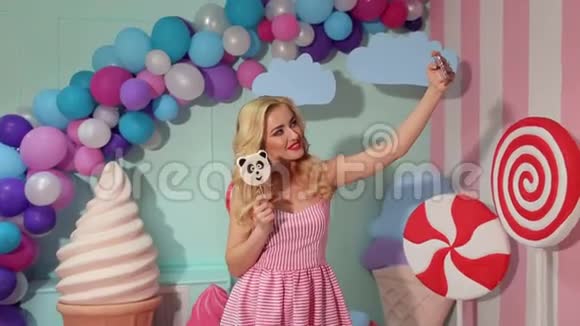 一个漂亮的年轻金发女郎穿着粉红色的裙子用一个大棒棒糖自拍视频的预览图