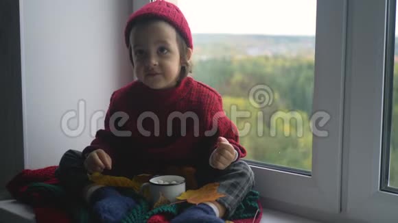 穿着红色毛衣和针织帽子的男孩坐在白色的大窗户上视频的预览图