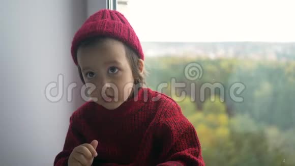 穿着红色毛衣和针织帽子的男孩坐在白色的大窗户上视频的预览图