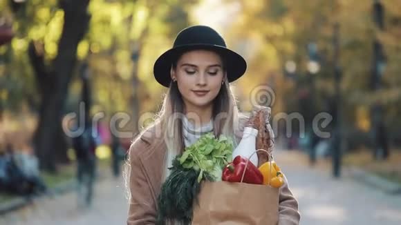 年轻漂亮的女人穿着时髦的外套在公园里散步拿着一包产品微笑着环顾四周视频的预览图