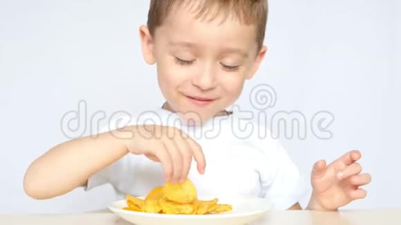 一个喜欢吃薯片的孩子坐在白色背景的桌子旁男孩正在吃薯片微笑着视频的预览图