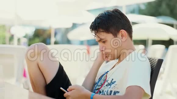 一个手里拿着智能手机的年轻人正坐在户外的桌子旁视频的预览图