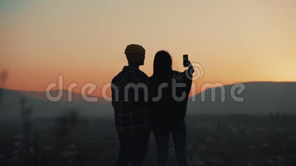 恋爱中的年轻夫妇在山上欣赏日落的剪影他们在智能手机上拍照假期视频的预览图
