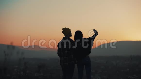恋爱中的年轻夫妇在山上欣赏日落的剪影他们在智能手机上拍照假期视频的预览图