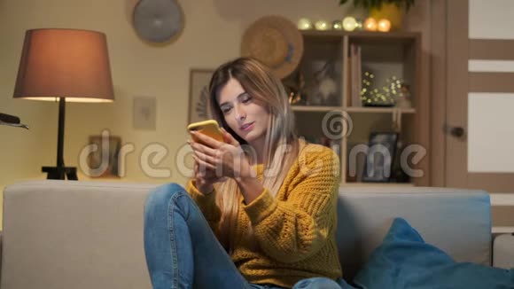 金发女郎黄色毛衣的肖像坐在沙发上回家在手机上聊天在手机上打字视频的预览图