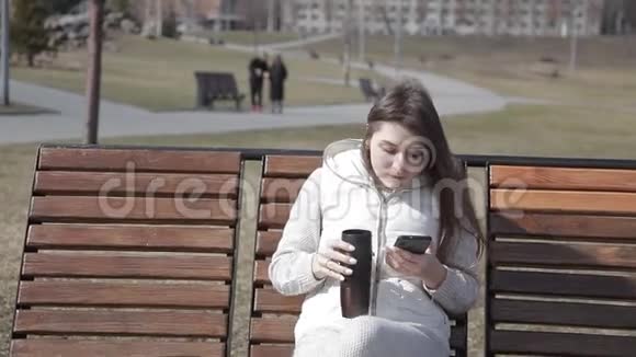 美丽的年轻女孩坐在城市公园的木凳上穿着白色西装喝着热杯咖啡视频的预览图