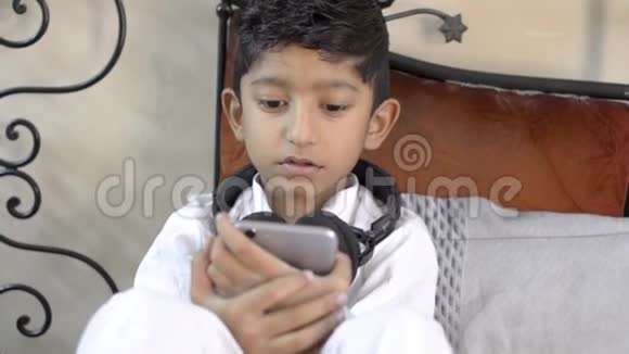 可爱的印度亚裔白人小男孩认真地看着手机屏幕脖子上戴着耳机的客厅clo视频的预览图
