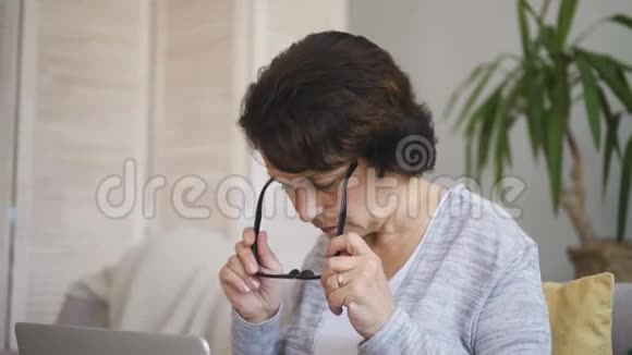 老年女性戴眼镜坐在公寓沙发上用笔记本电脑打字视频的预览图