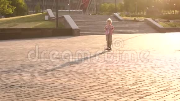 日落时分一个穿着红色格子衬衫的小男孩在城市公园骑滑板车视频的预览图