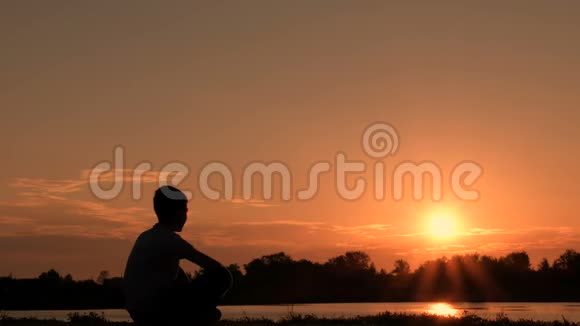 日出背景下少年的剪影那家伙坐在河岸上迎接黎明背光照明复制水疗中心视频的预览图