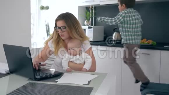 抑郁忙碌的妈妈手里拿着蹒跚学步的孩子在笔记本电脑后面工作背景是跳跃的儿子干扰视频的预览图