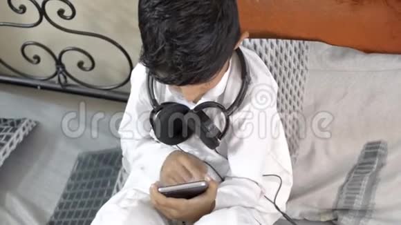 可爱的可爱的印度小白种人小孩在手机上忙碌智能手机屏幕上戴着耳机在卧室的脖子上视频的预览图