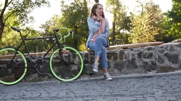 美丽的金发女孩坐在城市公园的长凳上她的徒步旅行自行车旁边她在说话视频的预览图
