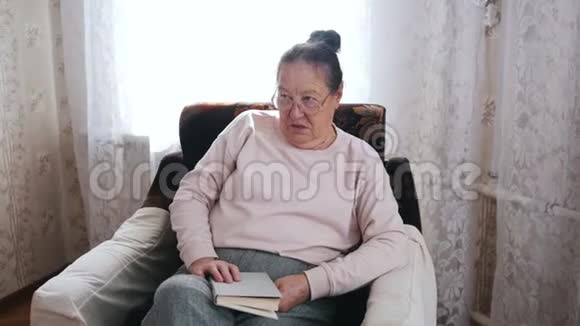 一位老妇人喜欢坐在椅子上在窗户的背景上大声朗读视频的预览图