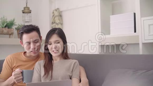 亚洲夫妇使用笔记本电脑在家里的客厅里喝着温暖的咖啡甜蜜的夫妇享受的时刻视频的预览图