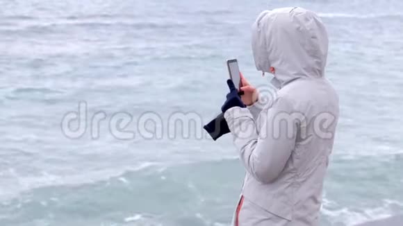 穿白色夹克的女人拿着手机大风浪后景视频的预览图