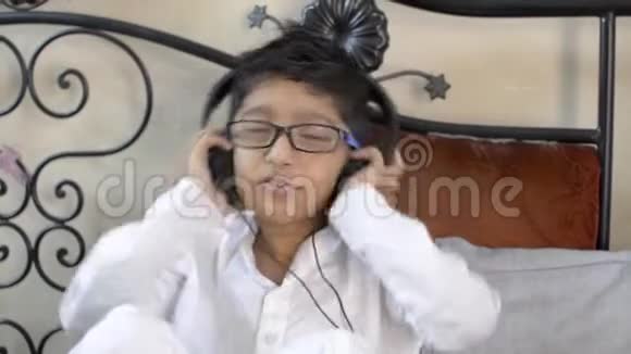 可爱的印度亚裔白种人小男孩戴着眼镜听着音乐戴着耳机跳舞点头坐在客厅里视频的预览图