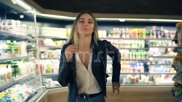 在超市快乐的年轻女孩在超市的货架之间滑稽地跳舞穿牛仔裤和黑色的金发女孩视频的预览图