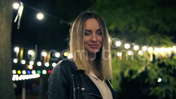 一个穿黑色皮夹克的漂亮女孩站在夜间公园的肖像咖啡馆的灯光五颜六色的灯视频的预览图